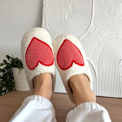 White slippers with red heart designs worn indoors against a textured white wall.