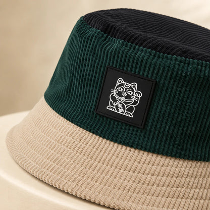 Josh & Tate - LuckyBrim™ Colorblock Bucket Hat - A close-up of a ribbed corduroy bucket hat with a dark green crown, a beige brim, and a black square patch featuring a white lucky cat illustration. 
