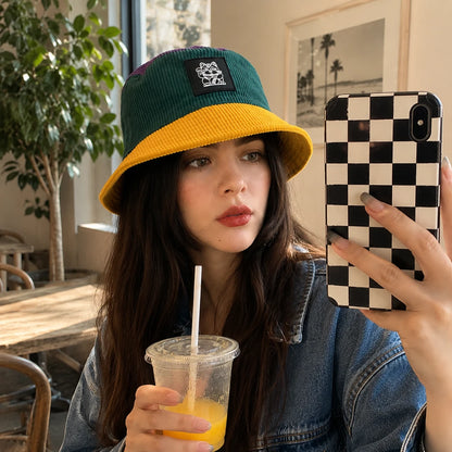 Josh & Tate - LuckyBrim™ Colorblock Bucket Hat - A woman takes a mirror selfie wearing a green and mustard yellow corduroy bucket hat with a black patch, holding an orange drink in one hand and a black-and-white checkered phone case in the other.