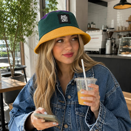 Josh & Tate - LuckyBrim™ Colorblock Bucket Hat - A woman in a cafe wearing a green and mustard yellow corduroy bucket hat with a black patch, holding an iced orange drink and a phone.