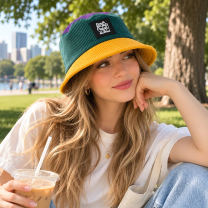 Josh & Tate - LuckyBrim™ Colorblock Bucket Hat - A woman sitting outdoors in a park wearing a corduroy bucket hat with a dark green crown, yellow brim, and purple side panels, along with a white t-shirt and light blue jeans, holding an iced coffee.