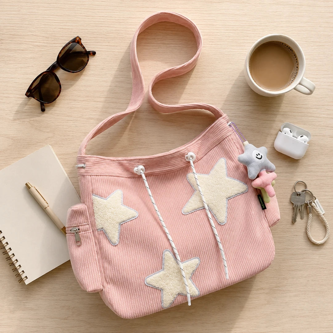 Josh & Tate - LuckyStar™ Ribbed Tote Bag - A pink corduroy shoulder bag with large cream star appliqués and white drawstring details, styled on a light wood surface with sunglasses, a notebook, pen, coffee cup, earbuds case, and keys nearby.