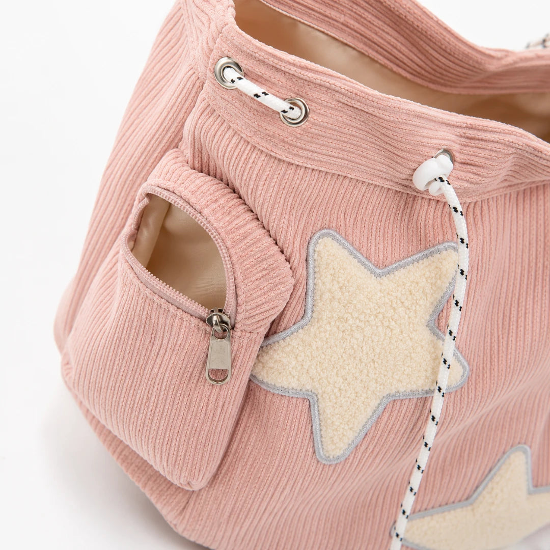 Josh & Tate - LuckyStar™ Ribbed Tote Bag  - A close-up of a dusty pink corduroy tote bag featuring a large cream-colored star applique with a gray outline on the front pocket, a zippered side pocket, and white drawstring cords with black speckles.