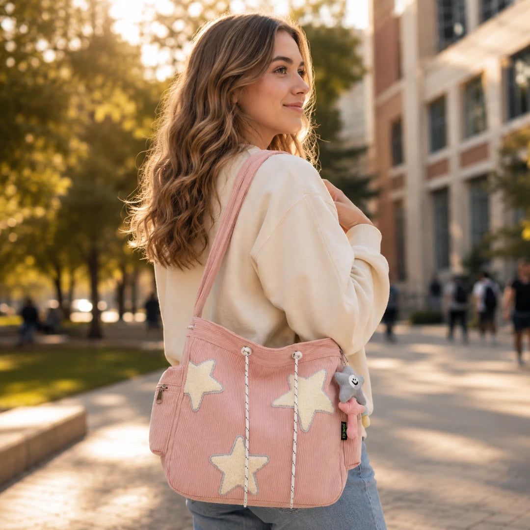 Josh & Tate - LuckyStar™ Ribbed Tote Bag  - A woman outdoors wearing a cream sweatshirt and jeans, carrying a pink canvas shoulder bag with large cream star appliqués and rope drawstrings. The background shows a softly blurred campus-like outdoor setting with trees and buildings.