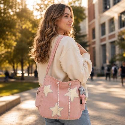 Josh & Tate - LuckyStar™ Ribbed Tote Bag  - A woman outdoors wearing a cream sweatshirt and jeans, carrying a pink canvas shoulder bag with large cream star appliqués and rope drawstrings. The background shows a softly blurred campus-like outdoor setting with trees and buildings.
