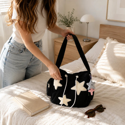 Josh & Tate - LuckyStar™ Ribbed Tote Bag  - A woman in a white t-shirt and light blue jeans holds a black velvet shoulder bag decorated with white and beige star appliqués, white drawstrings, and a small plush star charm, set in a bright bedroom.