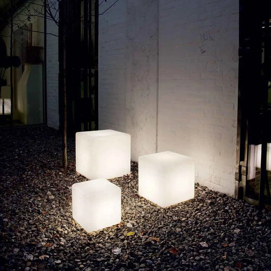 Three illuminated cube-shaped outdoor lights on a gravel surface against a brick wall.