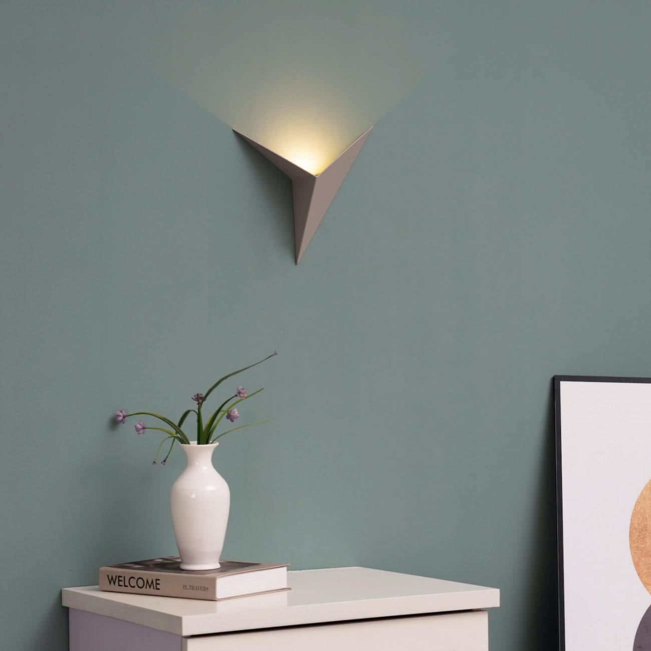 Wall-mounted lamp casting light on a table with a vase and books against a green wall.