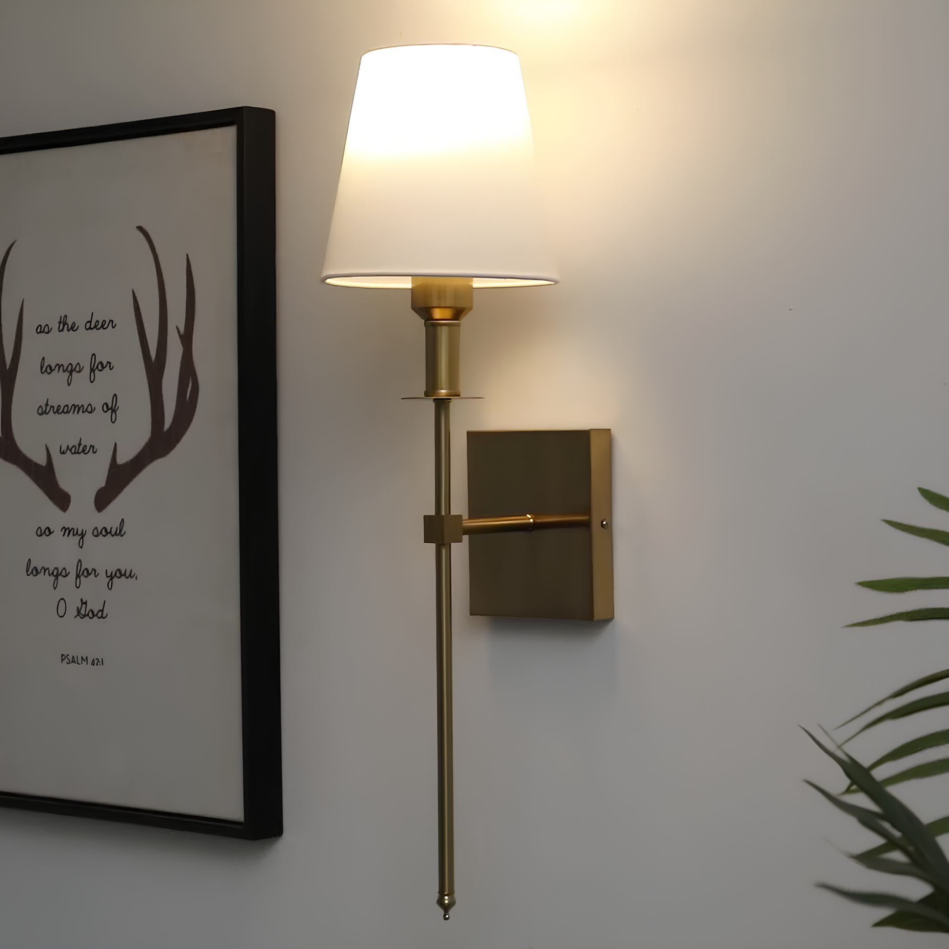 Wall-mounted lamp with a white shade and gold fixture on a light gray wall, next to a framed picture.