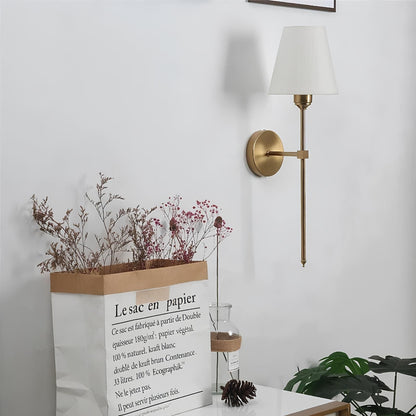 Gold wall sconce on a white wall with decorative elements like a plant and paper bag.