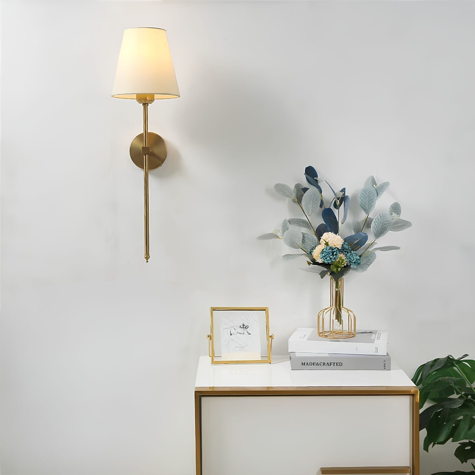 Decorative setup with a wall lamp, books, a vase with flowers, and a photo frame on a white surface.