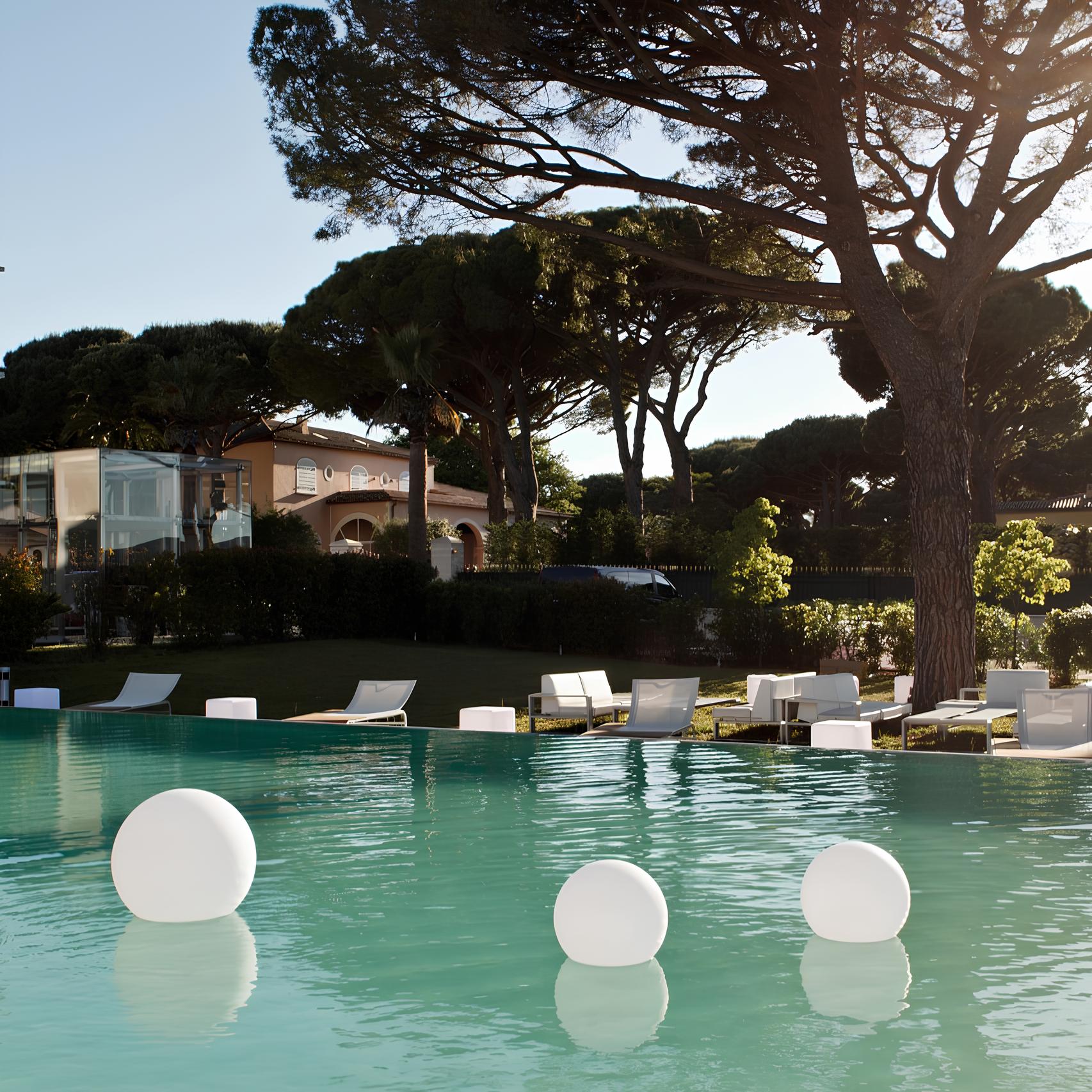 Pool area with lounge chairs and floating lights, surrounded by trees and buildings.