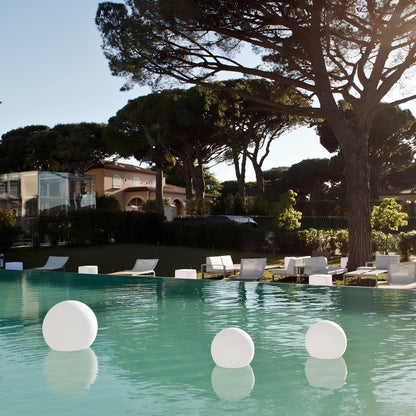 Pool area with lounge chairs and floating lights, surrounded by trees and buildings.
