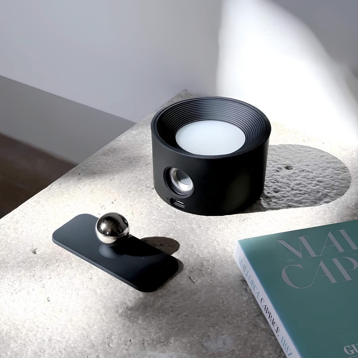 Black cylindrical object with a white candle inside on a concrete surface next to a book.