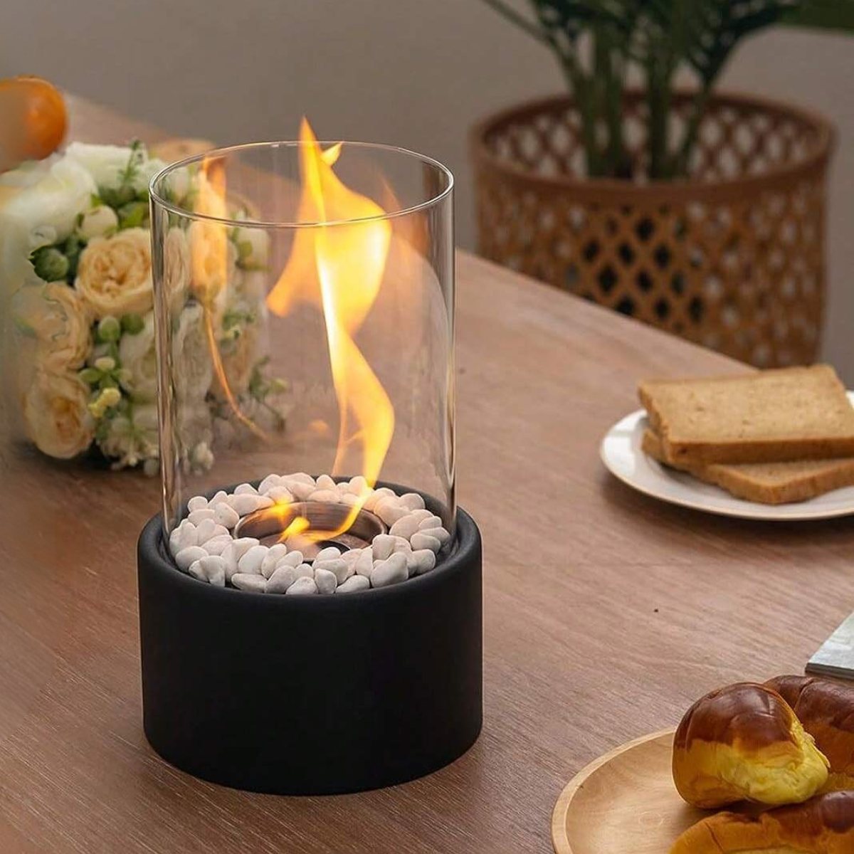 Tabletop fire pit with flames on a wooden table, surrounded by bread and flowers.