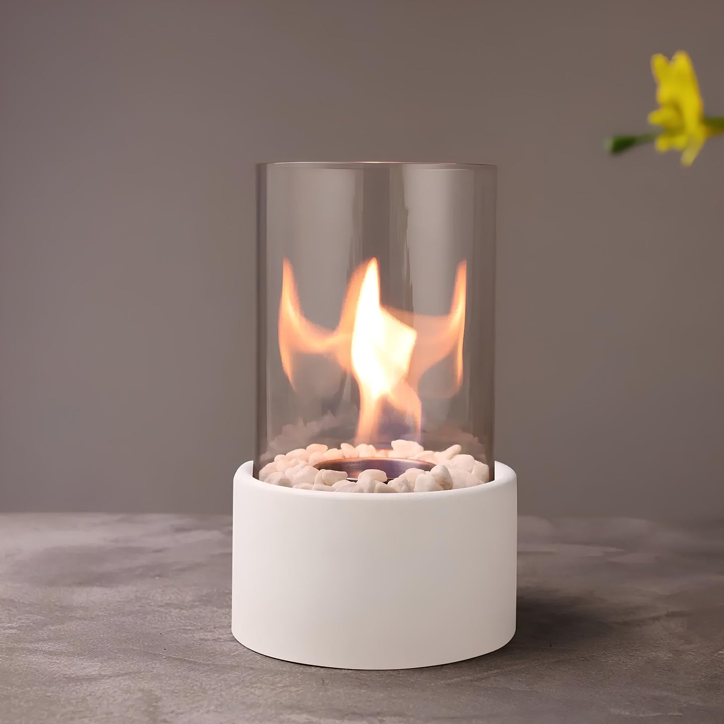 Small portable fireplace with glass cover on a gray surface, featuring a yellow flower in the corner.