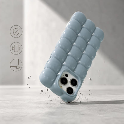 Josh & Tate - MarshBubble™ Puffy iPhone Case - A light blue textured phone case with a wavy, segmented bubble design, shown floating at an angle on a light gray surface with small debris around it. Three thin black feature icons appear on the left side of the image.