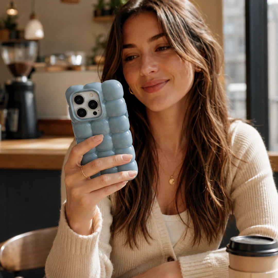 Josh & Tate - MarshBubble™ Puffy iPhone Case - A woman in a cream ribbed cardigan holds a light blue textured bubble phone case while looking at her phone in a warm, softly lit cafe setting.