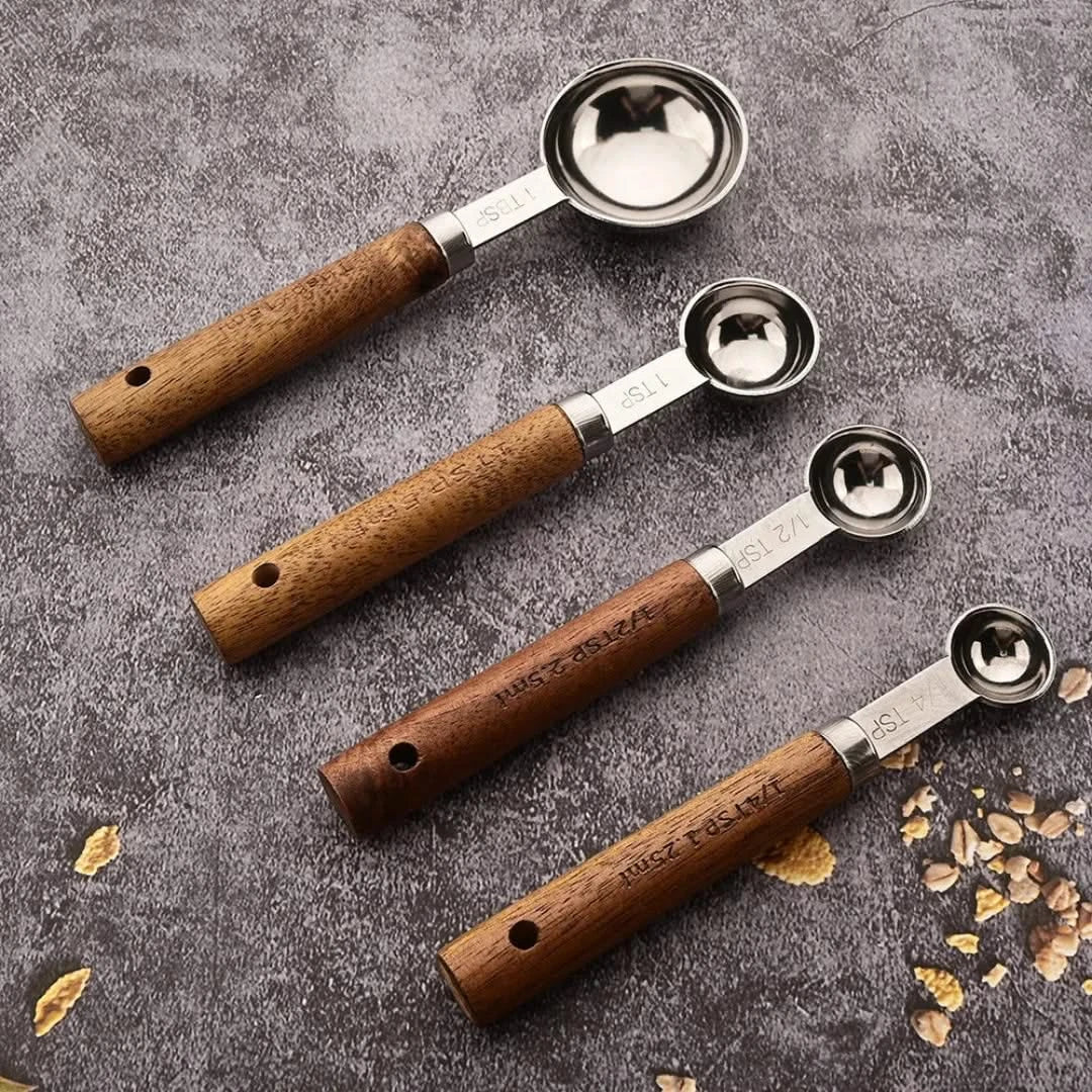 Josh & Tate - Measuring Spoon Set (Wood Handle) - Set of four wooden-handled metal measuring spoons on a textured surface with scattered grains.