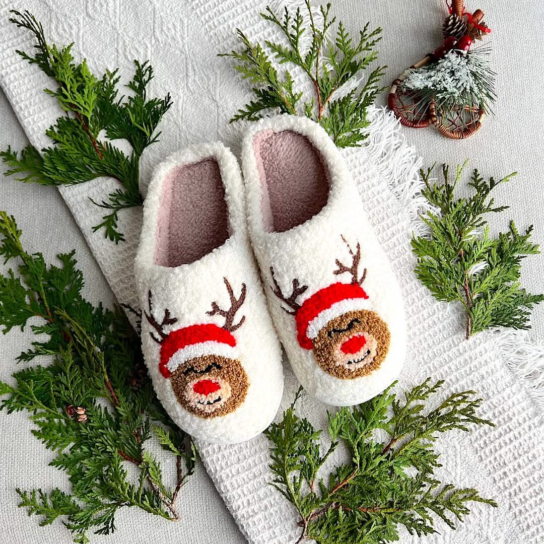 Pair of white slippers with reindeer faces on a textured surface with greenery.