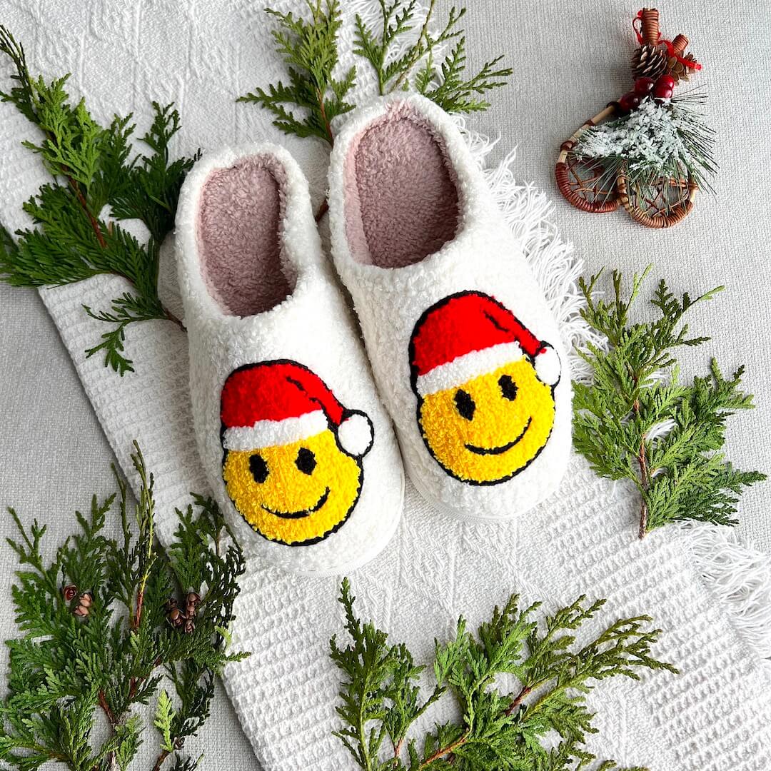 White fuzzy slippers with smiley face design wearing Santa hats on a textured surface with greenery.