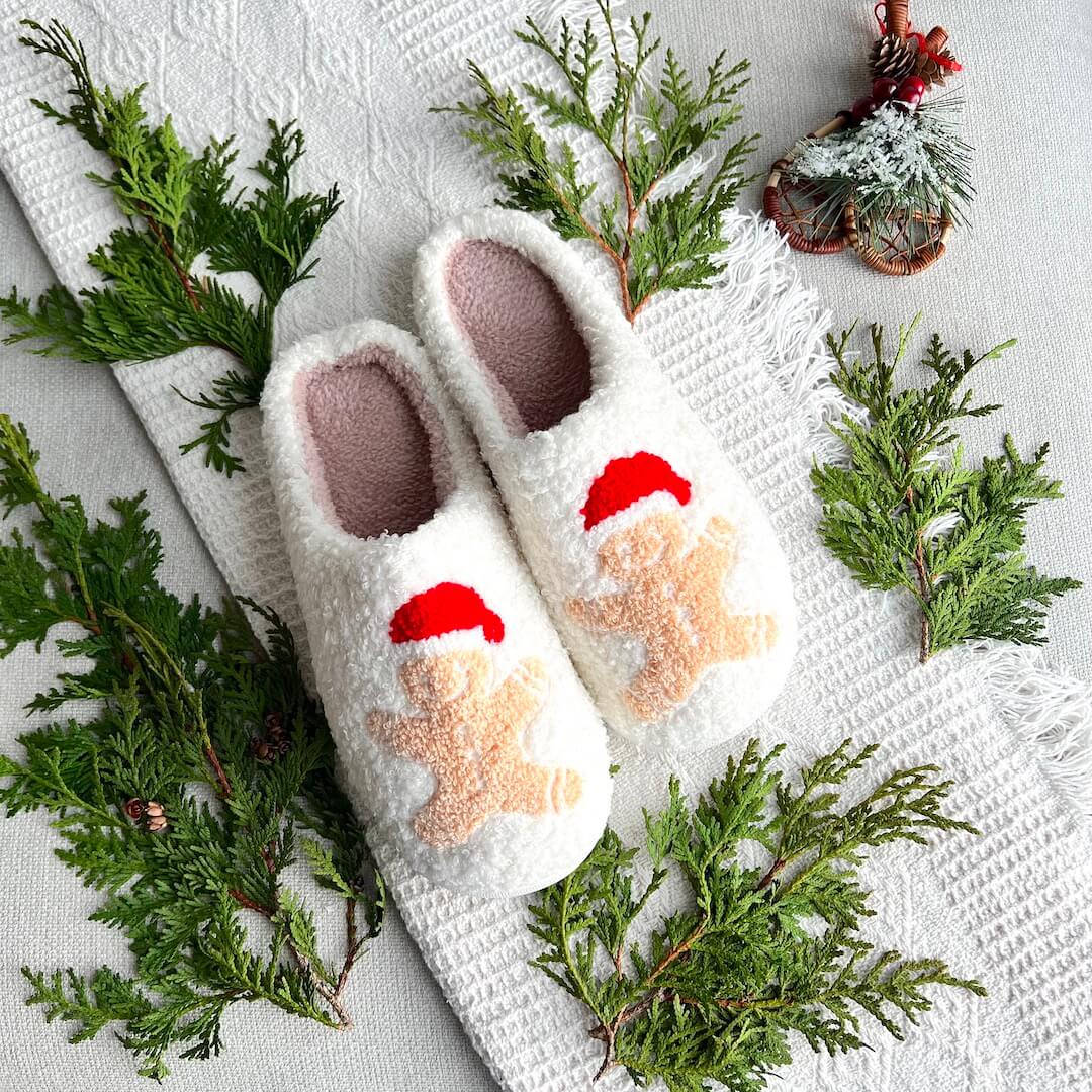 Pair of white slippers with gingerbread men design on a textured surface with greenery.