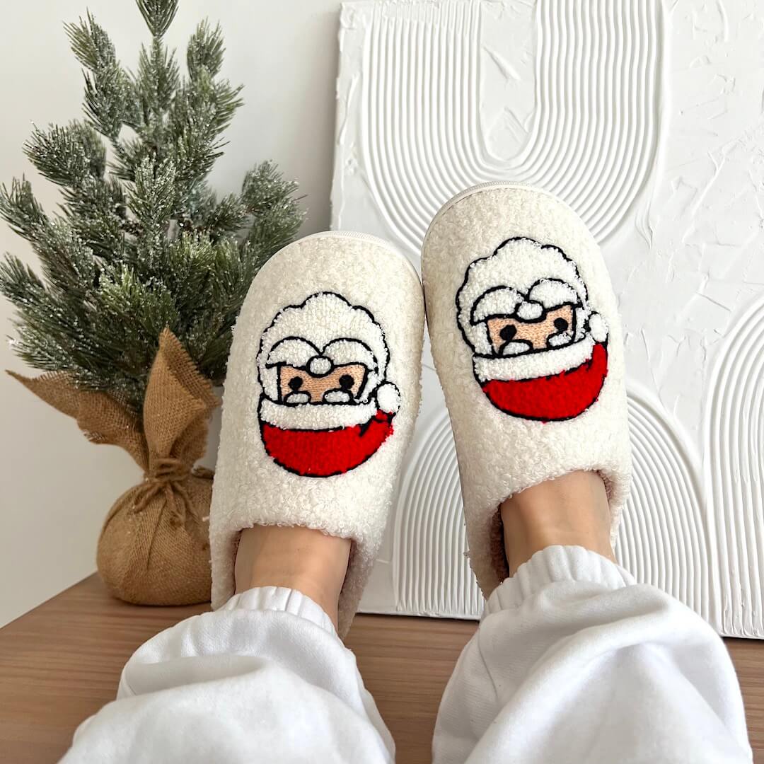 White fuzzy slippers with Santa Claus design worn indoors next to a small Christmas tree.