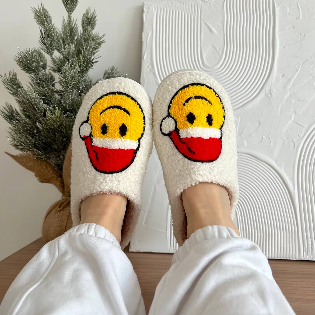 White fluffy slippers with emoji face design worn indoors, plant and textured wall in background