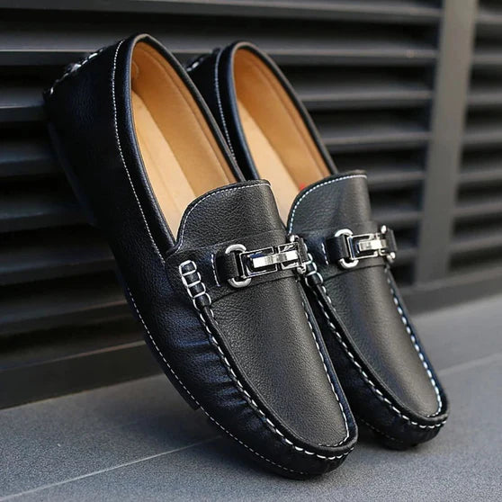 A pair of black Milano Classic Loafers with a sleek metal buckle on a city sidewalk.