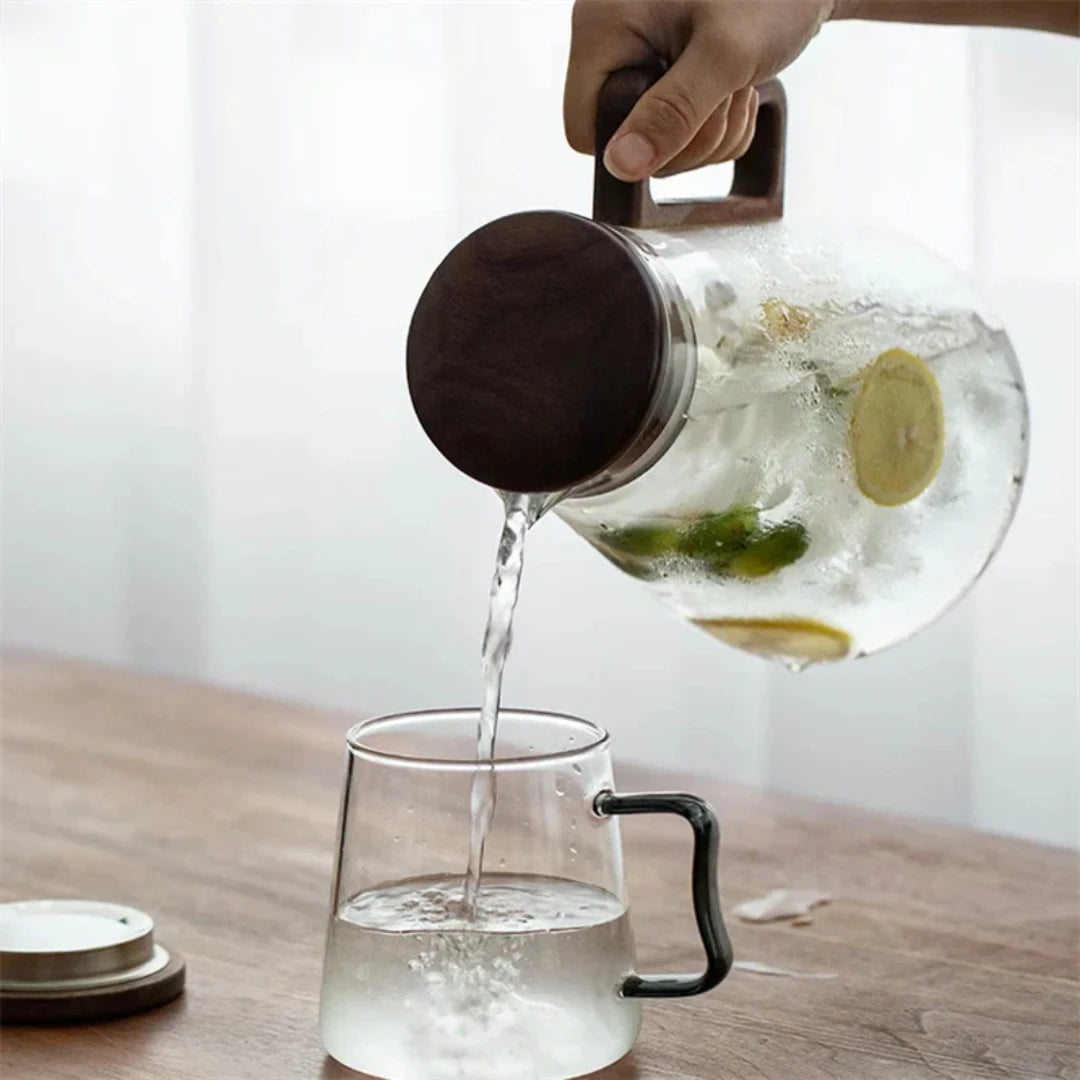 Josh & Tate - Montara Elegant Glass Pitcher - Person pouring tea from a glass teapot into a clear mug on a wooden surface.