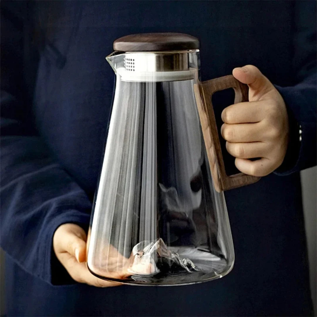 Josh & Tate - Montara Elegant Glass Pitcher - Glass teapot with wooden lid and handle held by a person against a dark background