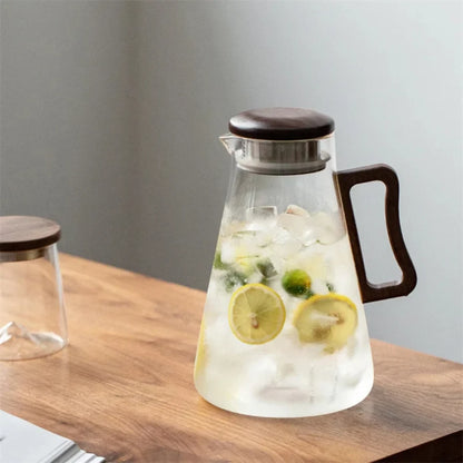Josh & Tate - Montara Elegant Glass Pitcher - Glass carafe with wooden lid and handle filled with iced water and lemon slices on a wooden surface.