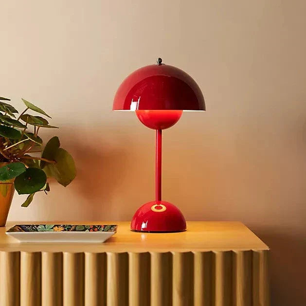 Red table lamp on a wooden surface with a plant and decorative plate in the background.