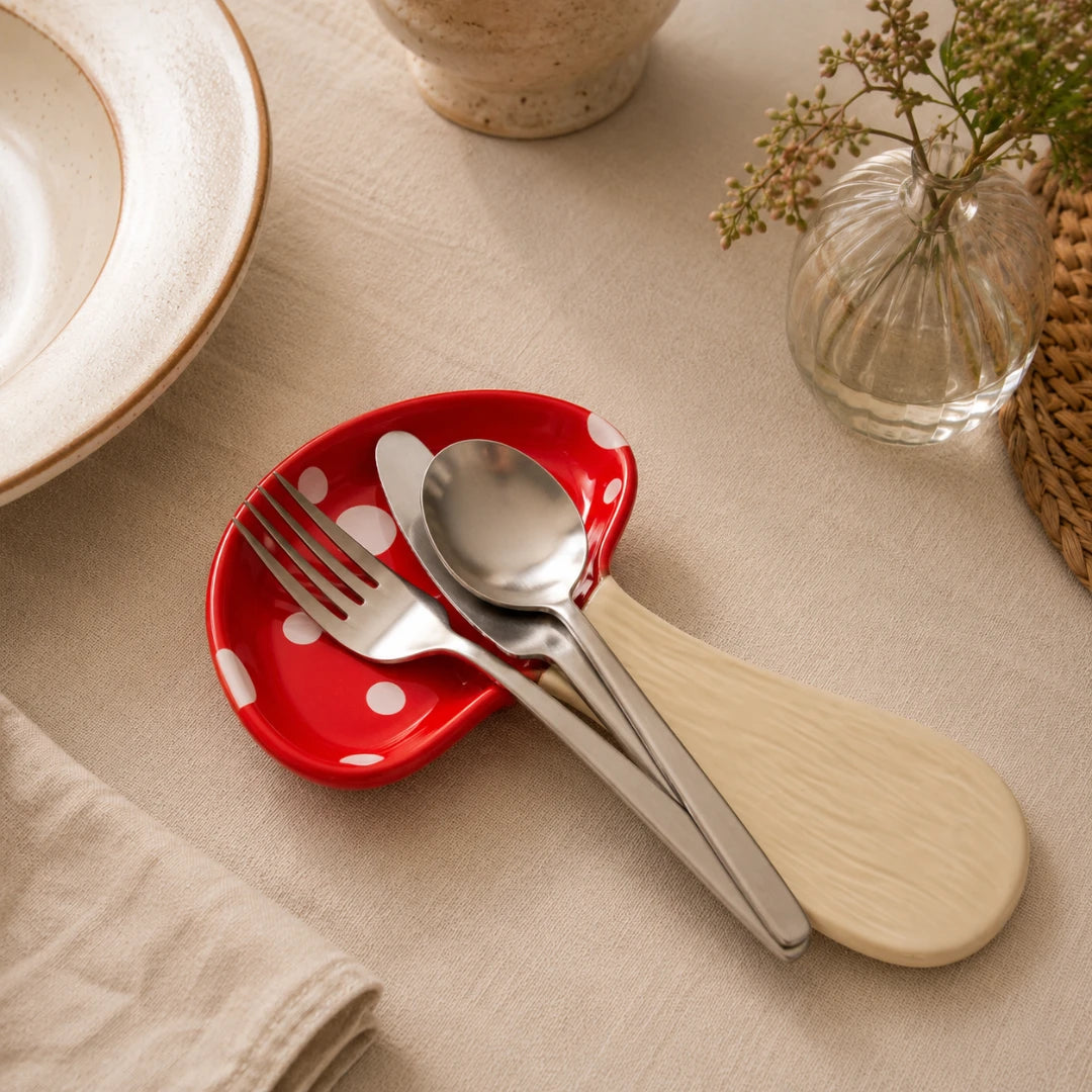 Josh & Tate - MushPerch™ Kitchen Spoon Holder - A red ceramic spoon rest with white polka dots holding silver metal cutlery, styled on a beige fabric table surface with a plate, vase, and woven mat nearby.
