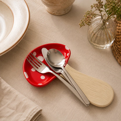 Josh & Tate - MushPerch™ Kitchen Spoon Holder - A red ceramic spoon rest with white polka dots holding silver metal cutlery, styled on a beige fabric table surface with a plate, vase, and woven mat nearby.
