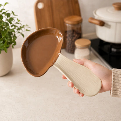 Josh & Tate - MushPerch™ Kitchen Spoon Holder - A hand holding a ceramic ladle with a brown bowl and a cream-colored handle, set against a blurred kitchen background.