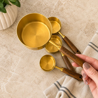 Josh & Tate - Natural Wood Measuring Cup Spoon Set - Set of gold measuring cups and spoons on a beige surface with a hand holding one spoon.