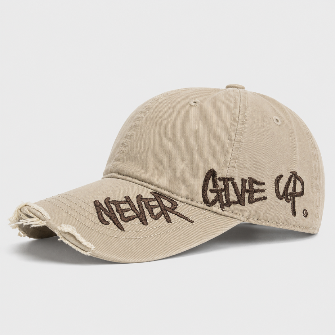 Josh & Tate - NeverGiveUp™ Baseball Cap - A beige distressed baseball cap with dark brown embroidered text reading 'NEVER GIVE UP' on the brim, shown against a light gray background.