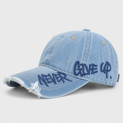Josh & Tate - NeverGiveUp™ Baseball Cap - A light blue denim baseball cap with distressed frayed edges and embroidered navy blue text reading 'NEVER GIVE UP.' on the brim, shown on a light gray background.