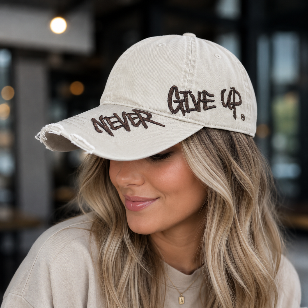 Josh & Tate - NeverGiveUp™ Baseball Cap - A woman wearing a distressed off-white baseball cap with dark brown embroidered text reading 'NEVER GIVE UP' on the front, photographed in a softly blurred indoor setting.