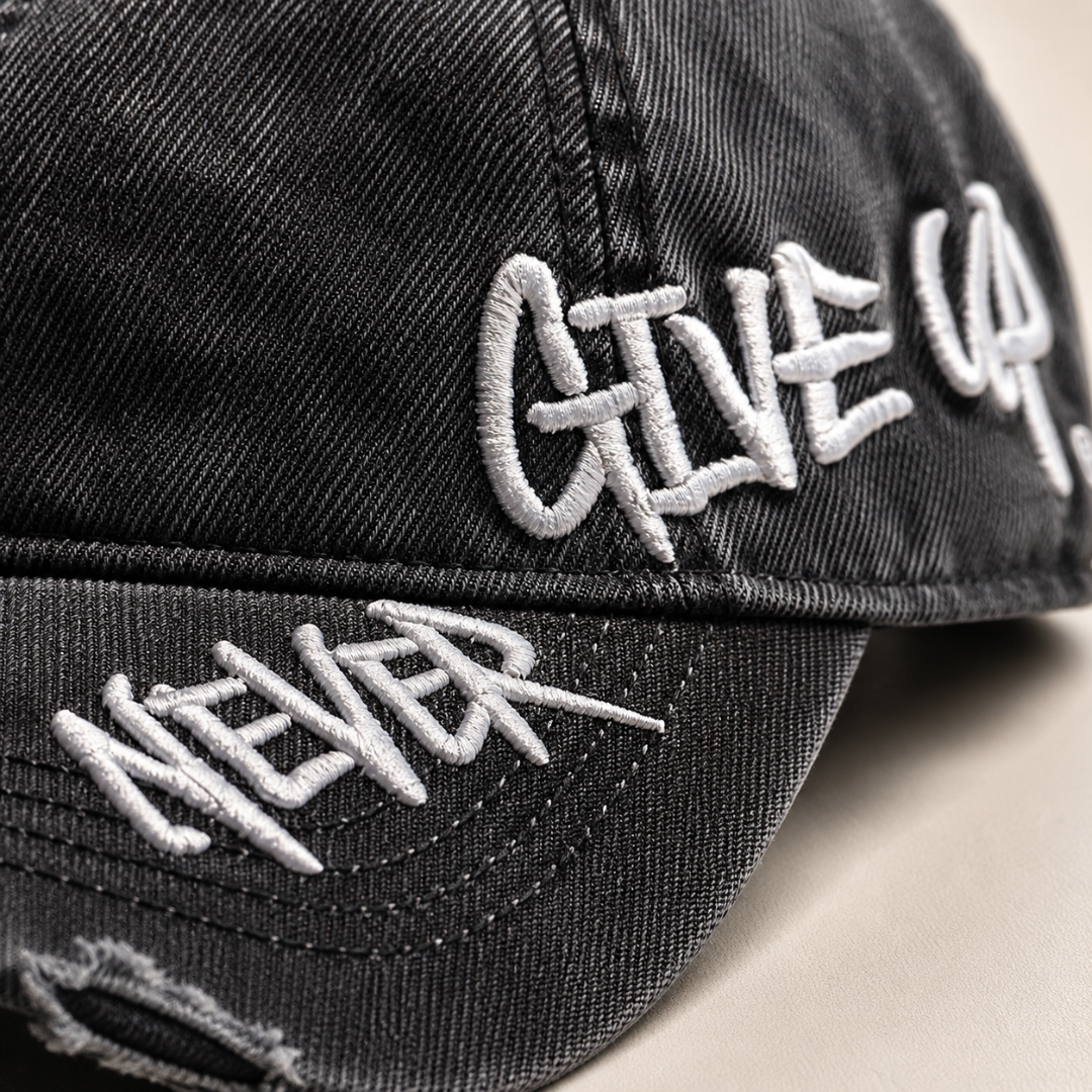 Josh & Tate - NeverGiveUp™ Baseball Cap - Close-up of a black distressed baseball cap with white embroidered text reading 'GIVE UP' on the side and 'NEVER' on the brim.