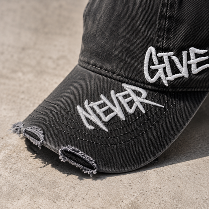 Josh & Tate - NeverGiveUp™ Baseball Cap - A close-up of a distressed black baseball cap with white embroidered text reading 'NEVER GIVE' on the brim, photographed outdoors on a light concrete surface.