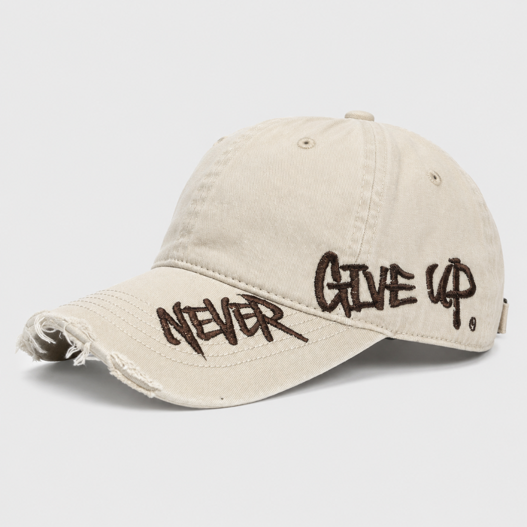 Josh & Tate - NeverGiveUp™ Baseball Cap - A distressed beige baseball cap with dark brown embroidered text reading 'NEVER GIVE UP' on the brim and side, shown on a light gray background.