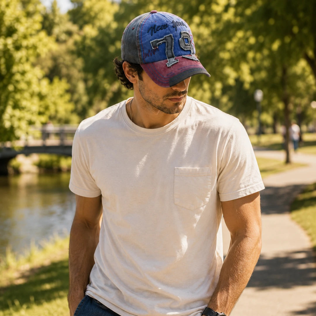 Josh & Tate - NewYork79™ Vintage Cap - A man wearing a white crewneck pocket t-shirt and a blue and purple distressed baseball cap with 'New Orleans 76ers' text, standing outdoors on a park path next to a river.