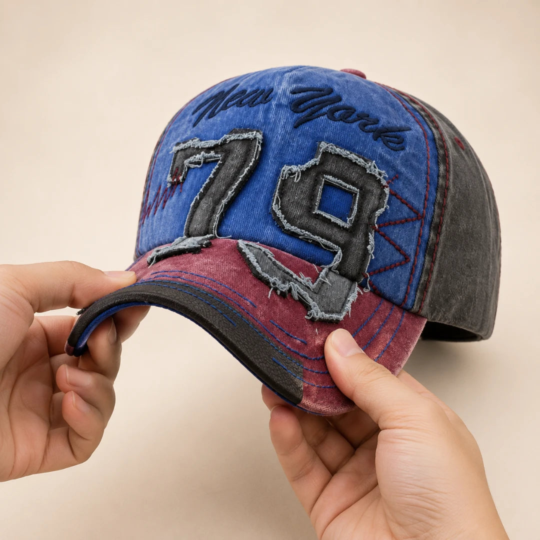Josh & Tate - NewYork79™ Vintage Cap - A distressed denim baseball cap being held by two hands against a plain beige background. The cap features a blue front panel with 'New York' embroidered in black script and a large '79' patch, a dark gray side panel, and a maroon brim.