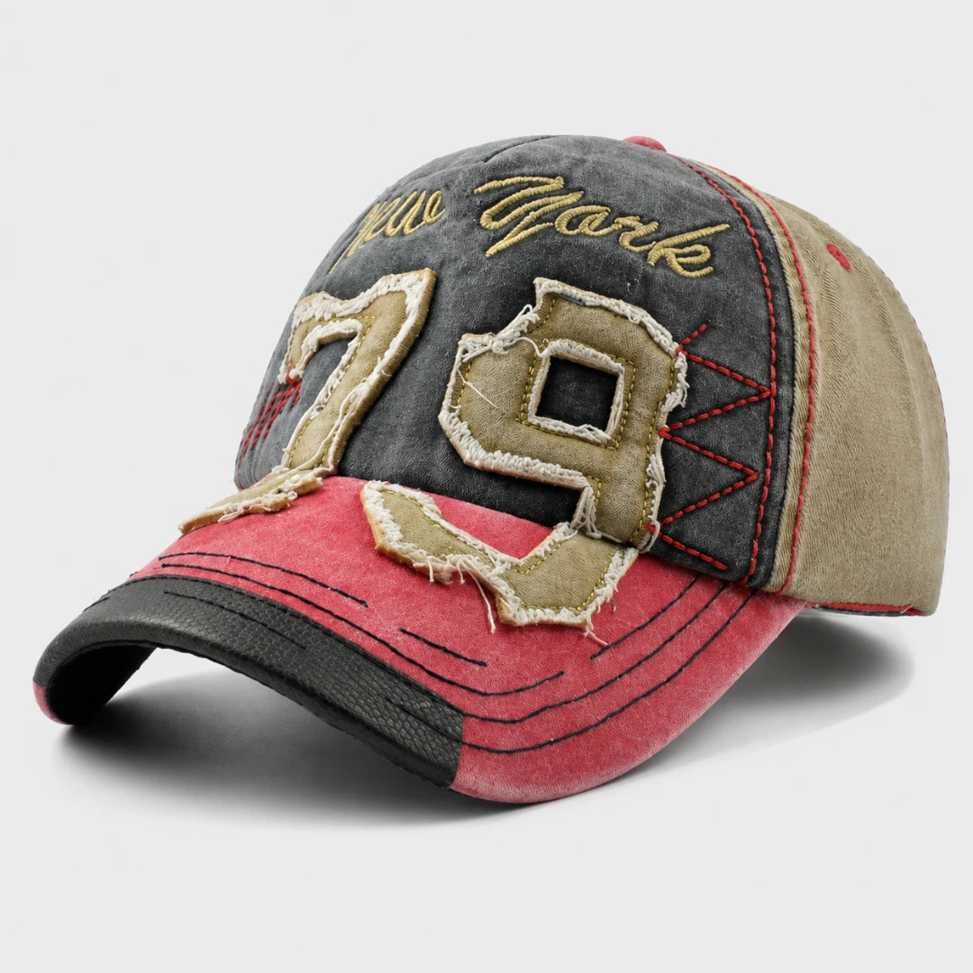 Josh & Tate - NewYork79™ Vintage Cap - A distressed baseball cap featuring a dark gray front panel with gold 'New York' script and large beige '79' patchwork numbers, a pink brim with black stitching, and beige side panels.