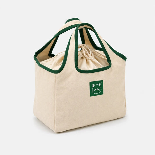 Josh & Tate - NookCarry™ Drawstring Lunch Tote - A beige canvas drawstring lunch bag with dark green trim on the handles and edges, featuring a green square patch with a mountain and bear logo on the front, set against a plain light gray background.