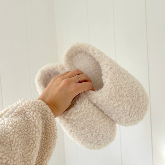 Beige fluffy slippers being held against a white wooden background