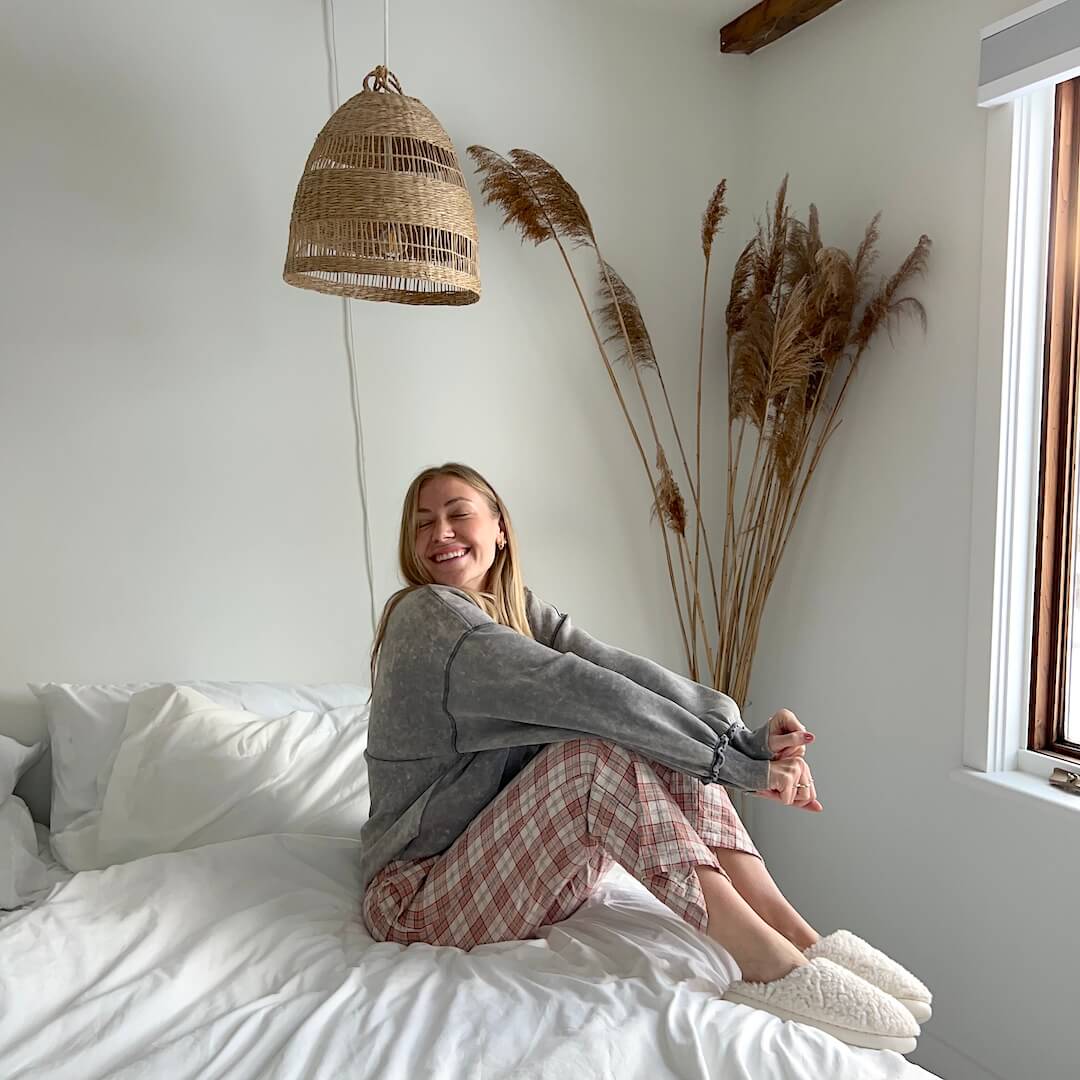 Woman sitting on a bed in a cozy room with a lamp and plants.