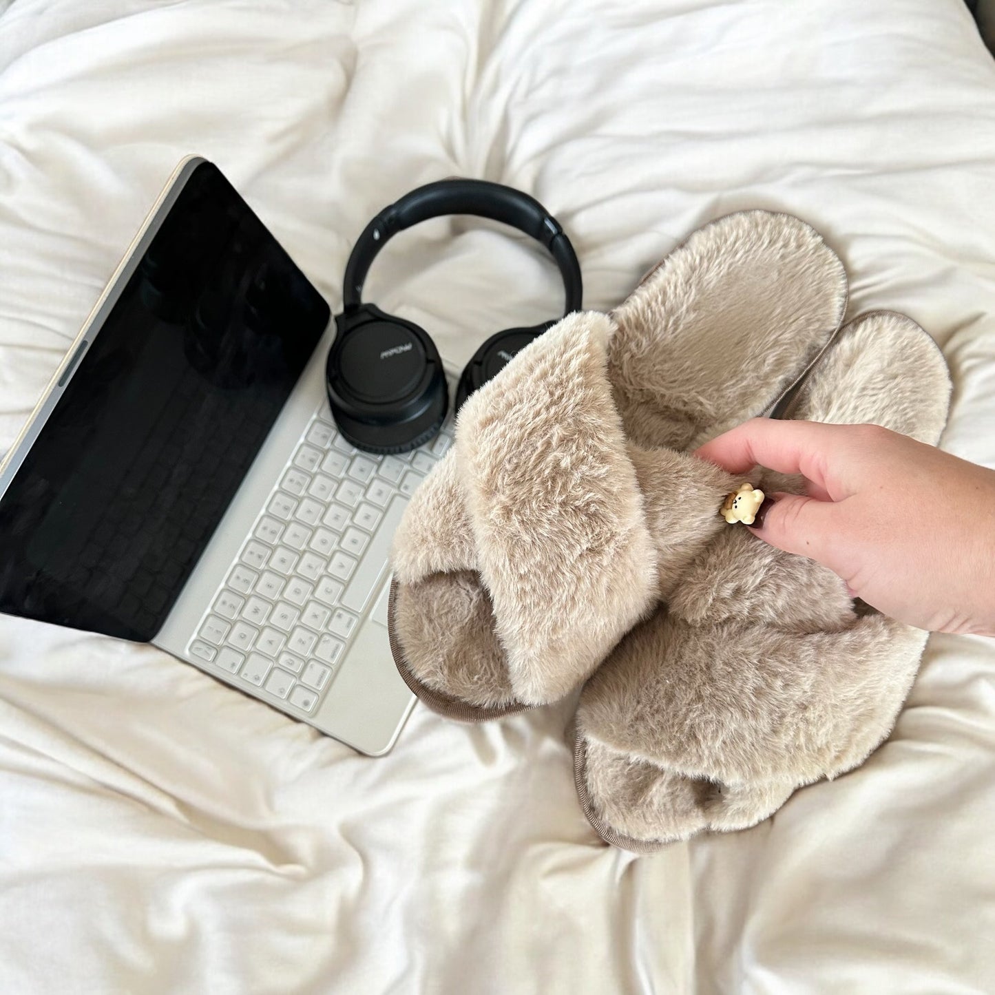 Beige slippers on a bed with a laptop, keyboard, and headphones.